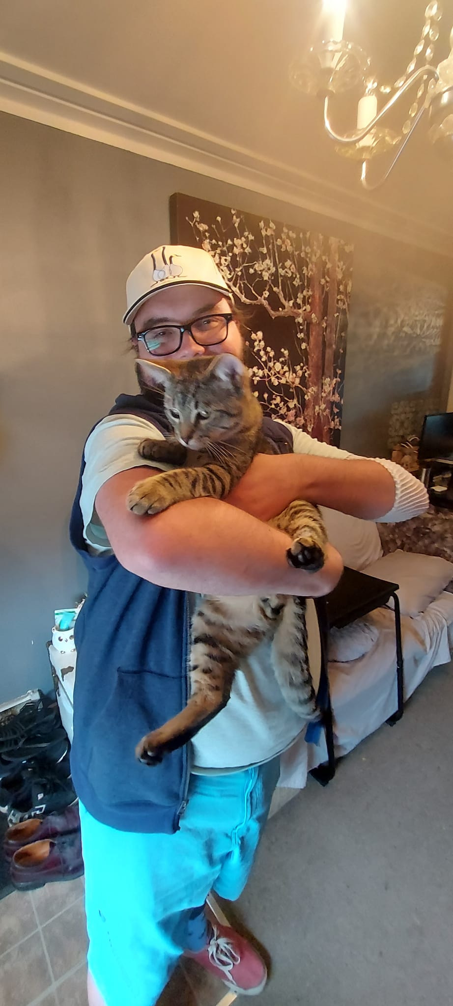 A man with a ball cap and bushy facial hair is holding a lanky brown tabby kitten, the kitten is squirming